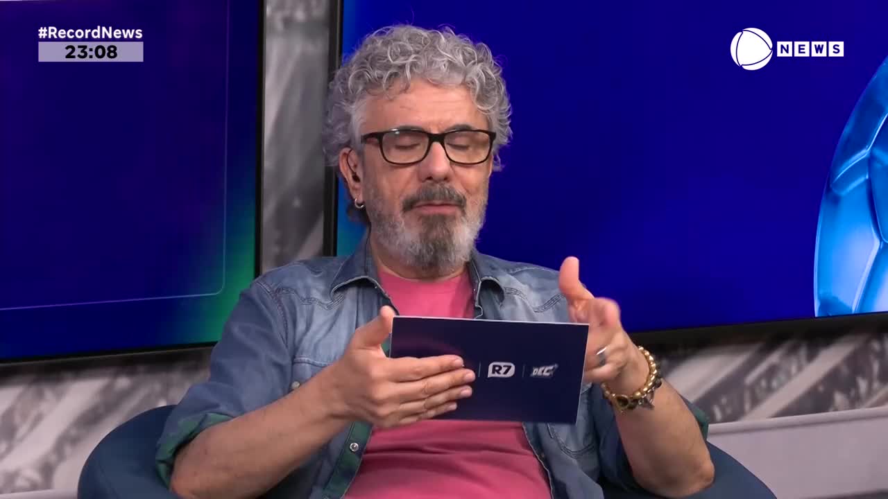 A man with curly gray hair and glasses gestures with his hands while holding a small blue tablet. The Record News logo appears on a screen behind him.