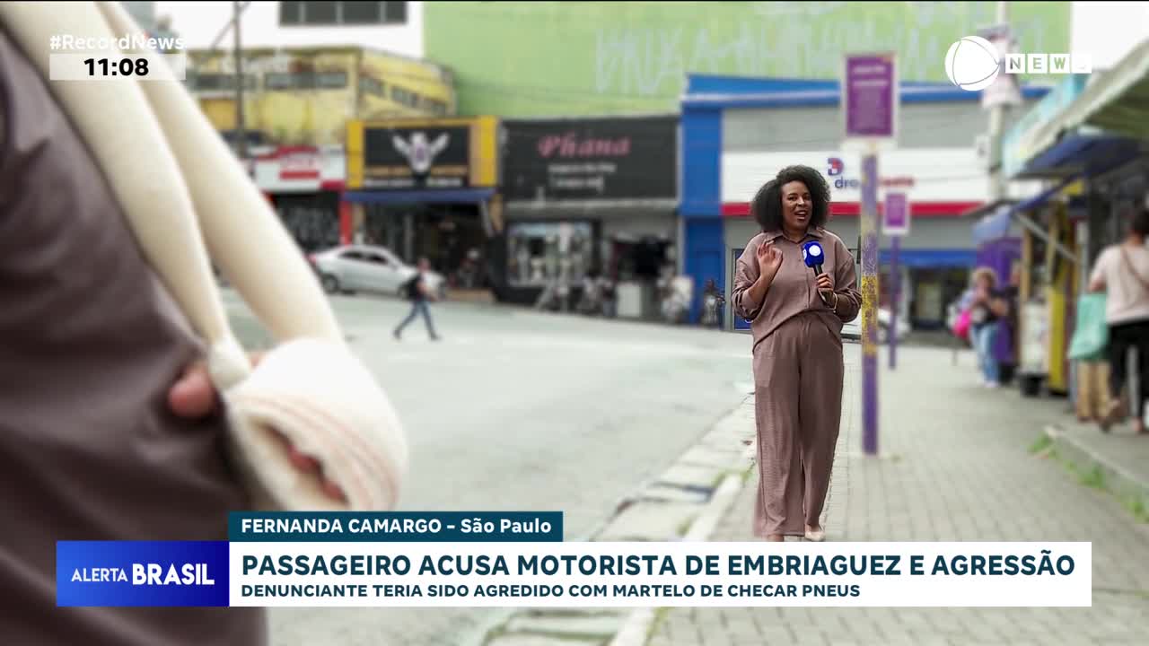 A reporter stands on a sidewalk in São Paulo, holding a microphone for Record News. In the foreground, a person's arm is bandaged and slung across their chest.