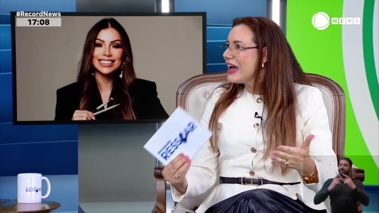 A woman in a white jacket gestures animatedly while holding a card. On a screen behind her, another woman smiles, holding what looks like scissors.