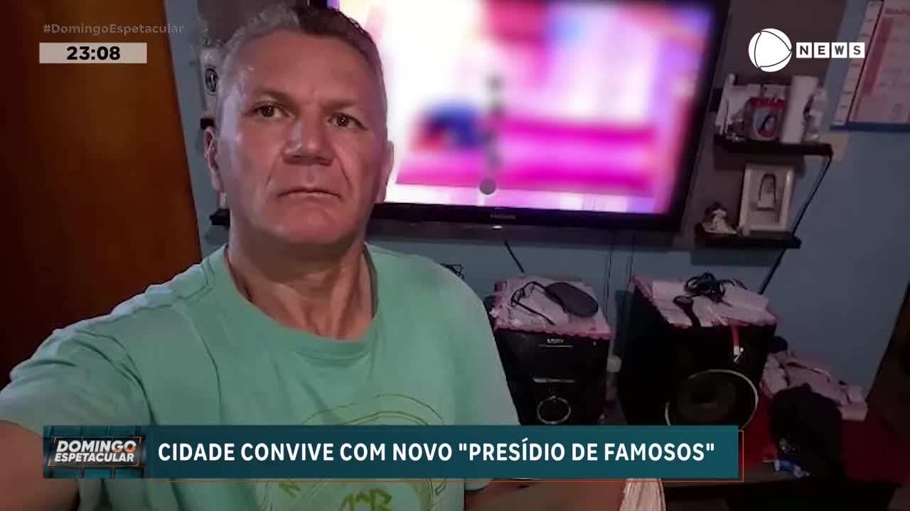 A man in a teal shirt looks directly at the camera, his expression unreadable. Behind him, a television screen displays a blurred image, and the Record News logo is visible in the corner.