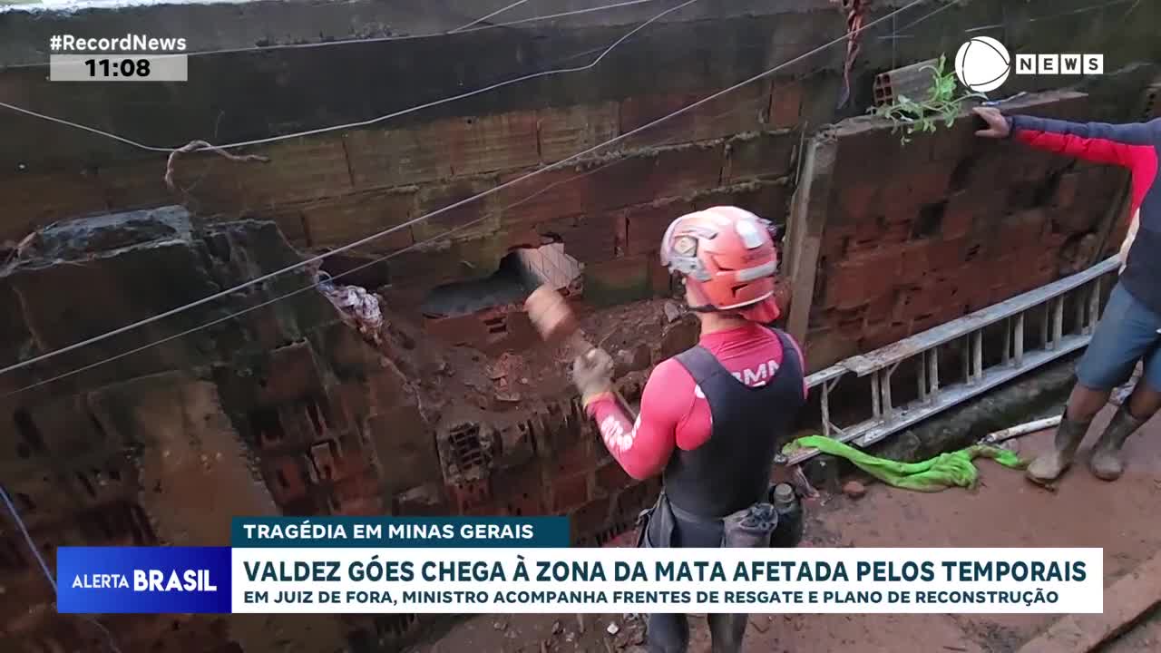 A rescuer in a red vest swings a sledgehammer at a collapsed brick wall. Another person stands nearby, observing the efforts in this area of Minas Gerais.
