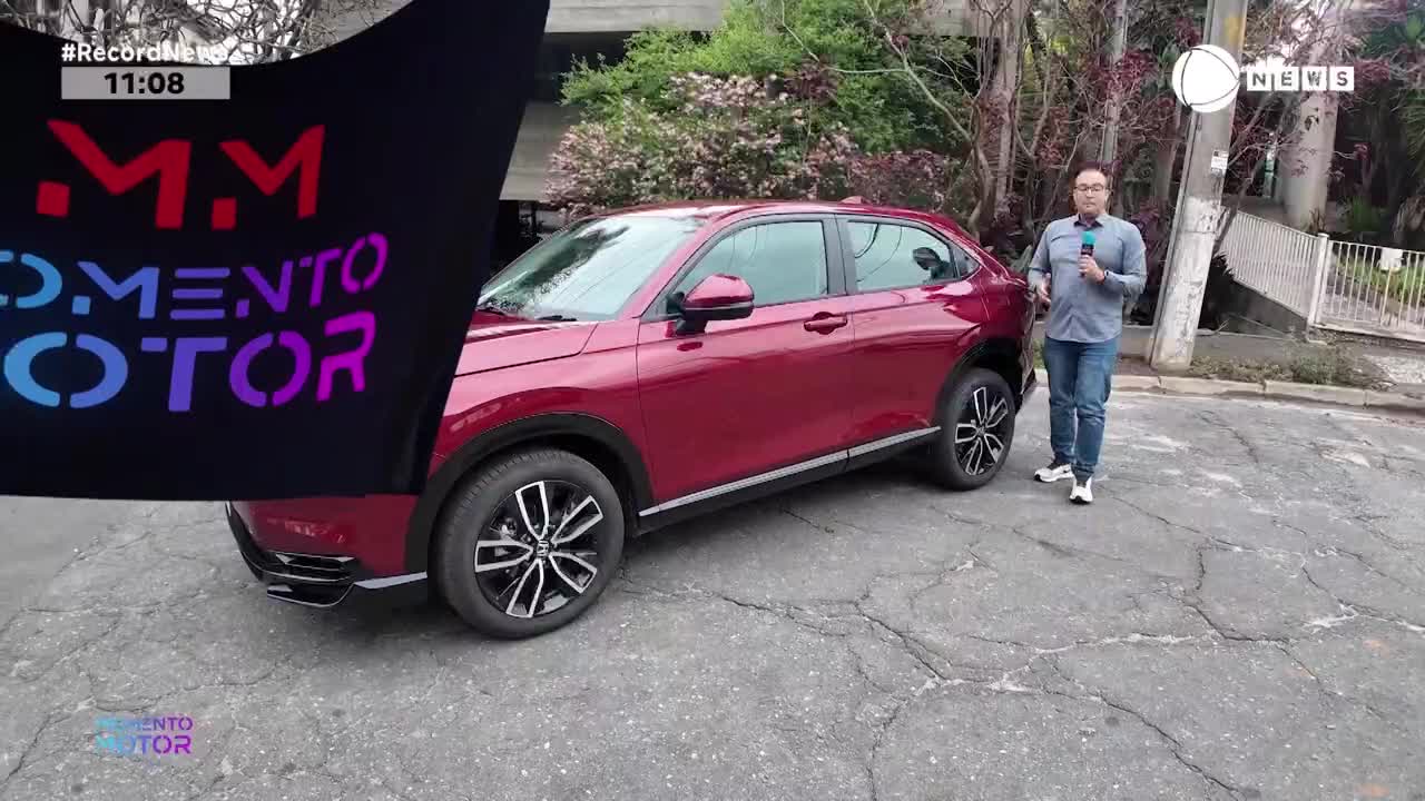 A dark banner with "Momento Motor" emblazoned in colorful letters is being pulled aside, revealing a shiny red SUV. A man in a blue shirt and jeans stands beside the car, holding a microphone, ready to speak for Record News.