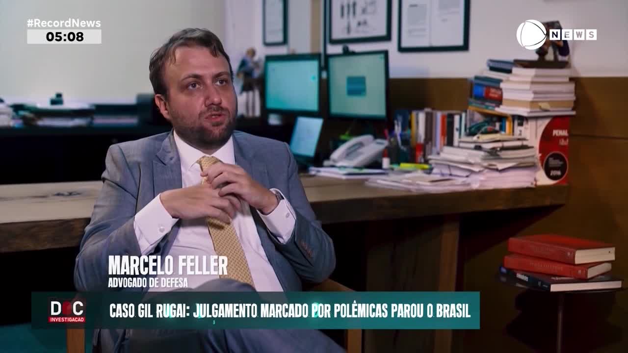 A man in a suit adjusts his tie while speaking. Behind him, a desk overflows with papers and books, a typical scene for a legal professional in Brazil.
