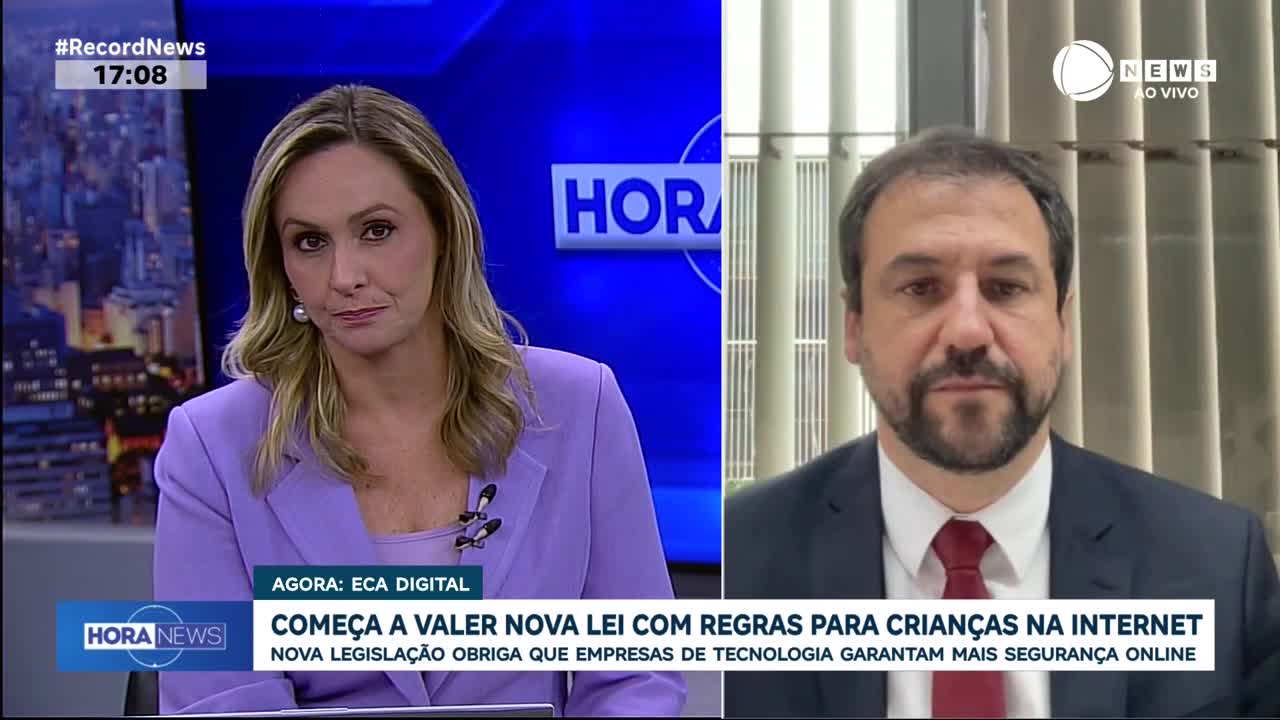 A woman in a lavender jacket sits in front of a blue screen displaying the Record News logo and the time, 17:08. To her right, a man in a suit and red tie speaks on a video call, his background a wall of vertical blinds.