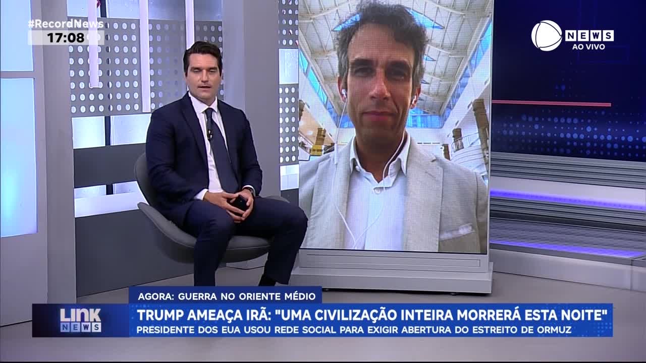 A man in a dark suit sits on a chair, hands clasped, facing the camera. To his right, a large screen displays a man in a light jacket, speaking with earphones on. A man in a dark suit sits on a chair, hands clasped, facing the camera. To his right, a large screen displays a man in a light jacket, speaking with earphones on.