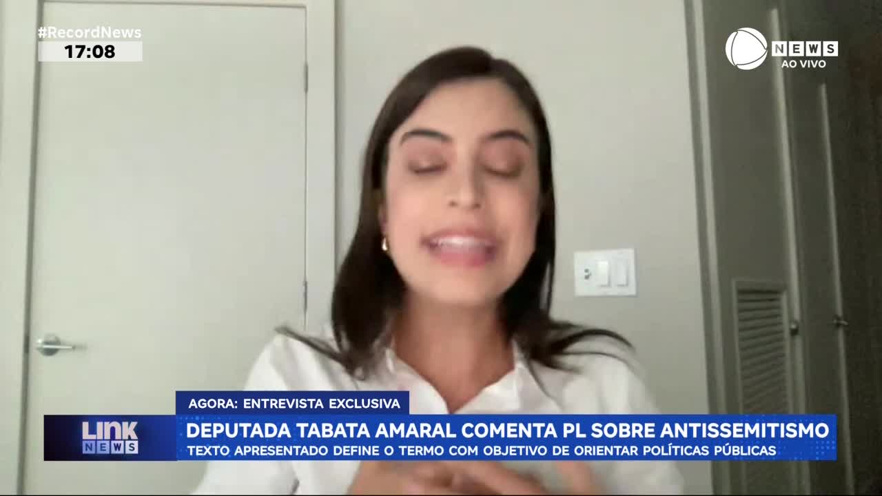 A young woman with dark hair speaks animatedly, her mouth open mid-sentence. A Record News chyron identifies her as Deputada Tabata Amaral, discussing a bill about antisemitism. A young woman with dark hair speaks animatedly, her mouth open mid-sentence. A Record News chyron identifies her as Deputada Tabata Amaral, discussing a bill about antisemitism.