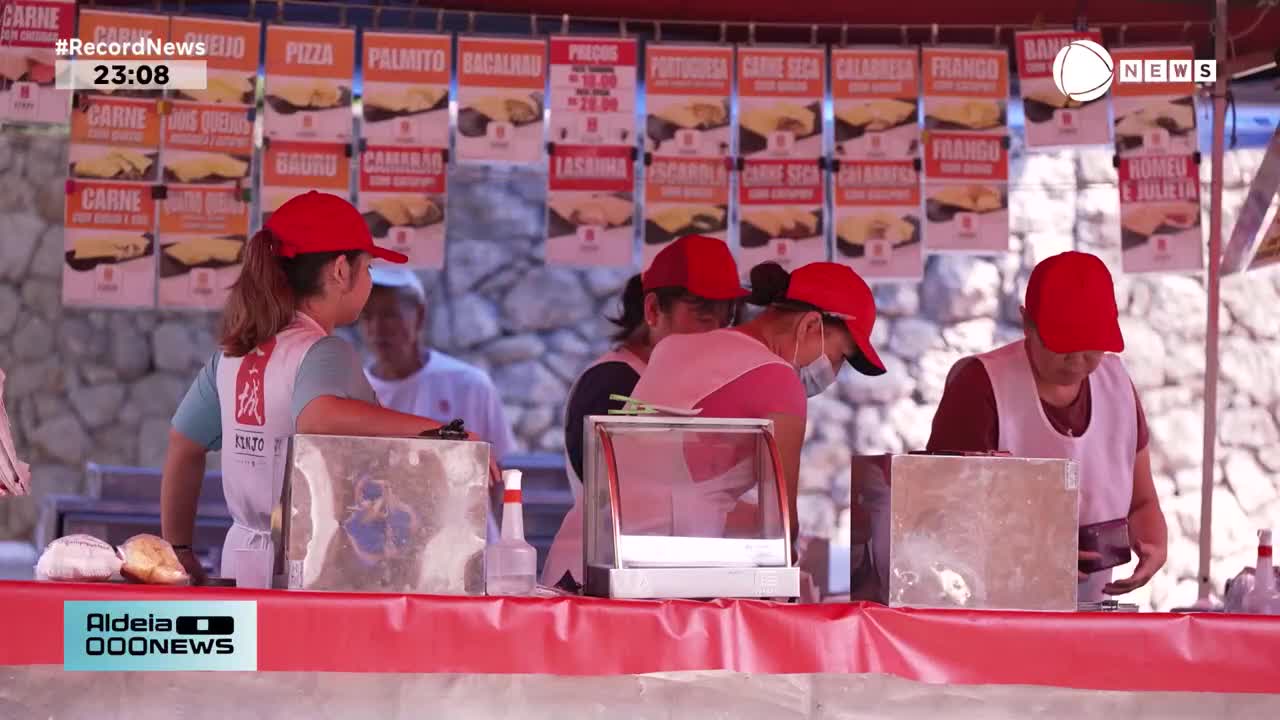 Vendors in red caps work behind a food stall, arranging items under a canopy displaying menu signs. A man in a white shirt stands behind them, watching the activity. Vendors in red caps work behind a food stall, arranging items under a canopy displaying menu signs. A man in a white shirt stands behind them, watching the activity.