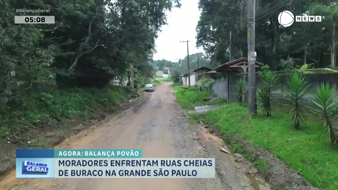 A white car slowly navigates a muddy, unpaved road lined with trees and simple houses. The road is deeply rutted, with puddles and debris scattered along its edges. A white car slowly navigates a muddy, unpaved road lined with trees and simple houses. The road is deeply rutted, with puddles and debris scattered along its edges.