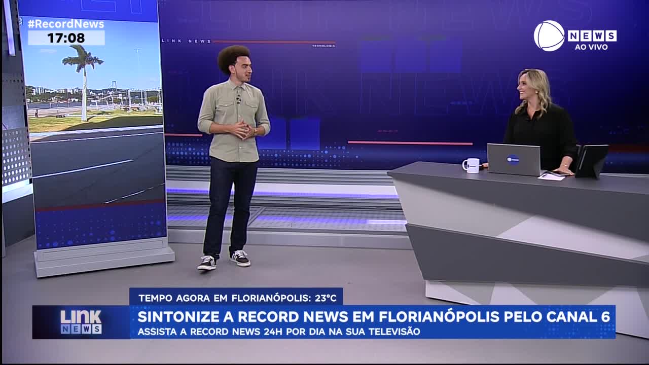 A man with an afro stands near a large screen displaying a tropical scene and the time. Across the studio, a woman smiles from behind a modern desk, a laptop open before her. A man with an afro stands near a large screen displaying a tropical scene and the time. Across the studio, a woman smiles from behind a modern desk, a laptop open before her.