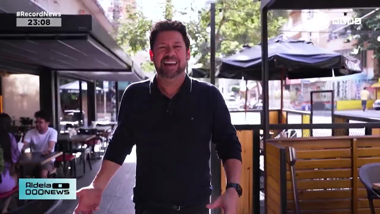 A man in a dark polo shirt laughs heartily in front of an outdoor cafe. Behind him, people sit at tables, and a lone figure walks down the street. A man in a dark polo shirt laughs heartily in front of an outdoor cafe. Behind him, people sit at tables, and a lone figure walks down the street.