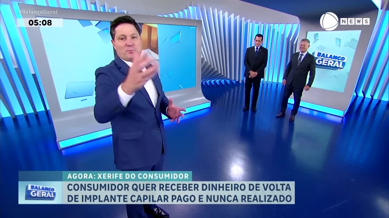 A man in a blue suit gestures animatedly in a bright studio. Two other men in suits stand behind him, facing the camera. A man in a blue suit gestures animatedly in a bright studio. Two other men in suits stand behind him, facing the camera.