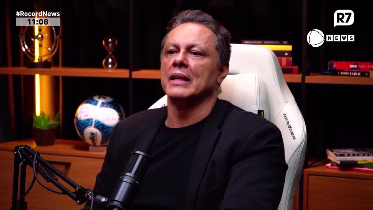 A man in a black jacket speaks into a microphone. Behind him, a shelf holds a soccer ball and other decorative items.