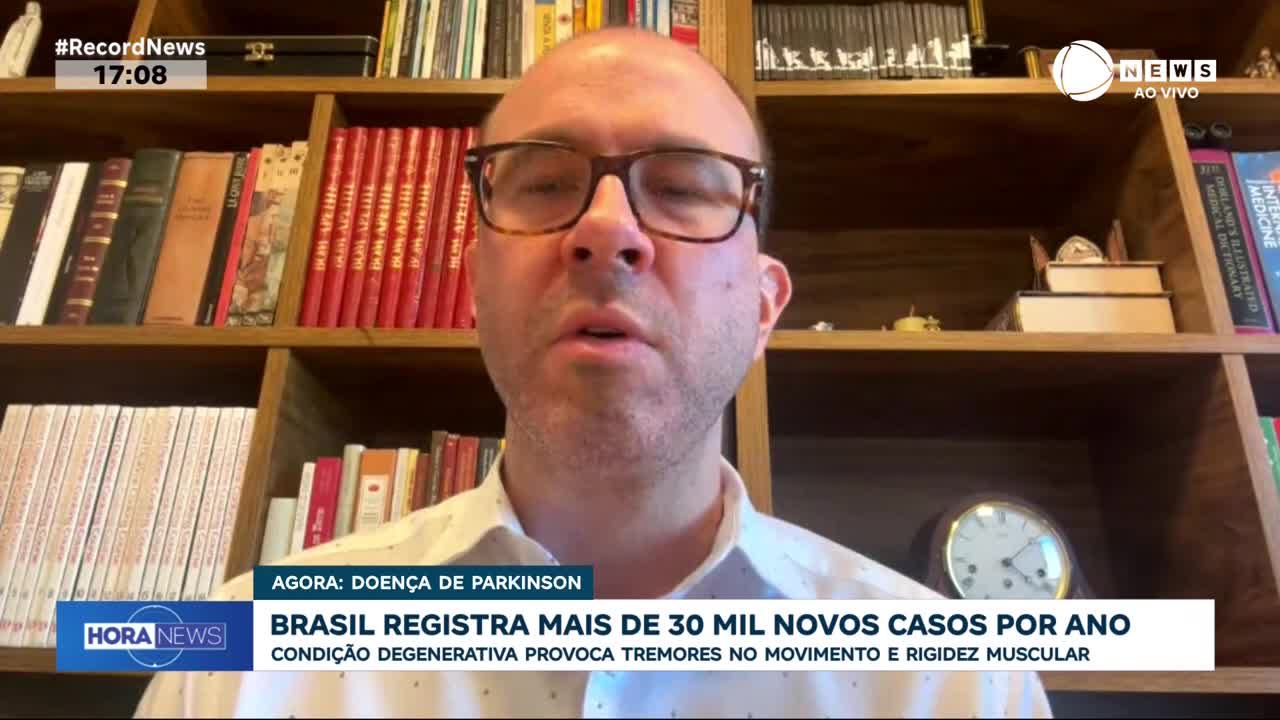A man with glasses speaks, framed by bookshelves filled with books. A news ticker at the bottom of the screen reports on Parkinson's disease cases in Brazil.