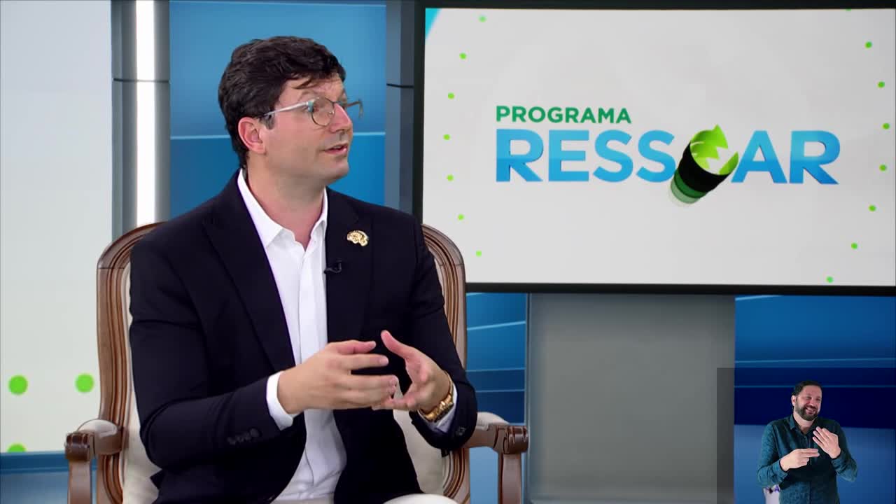 A man in a dark suit and white shirt gestures with his hands as he speaks. Behind him, a screen displays "PROGRAMA RESSAR" with a green, rolled-up graphic.