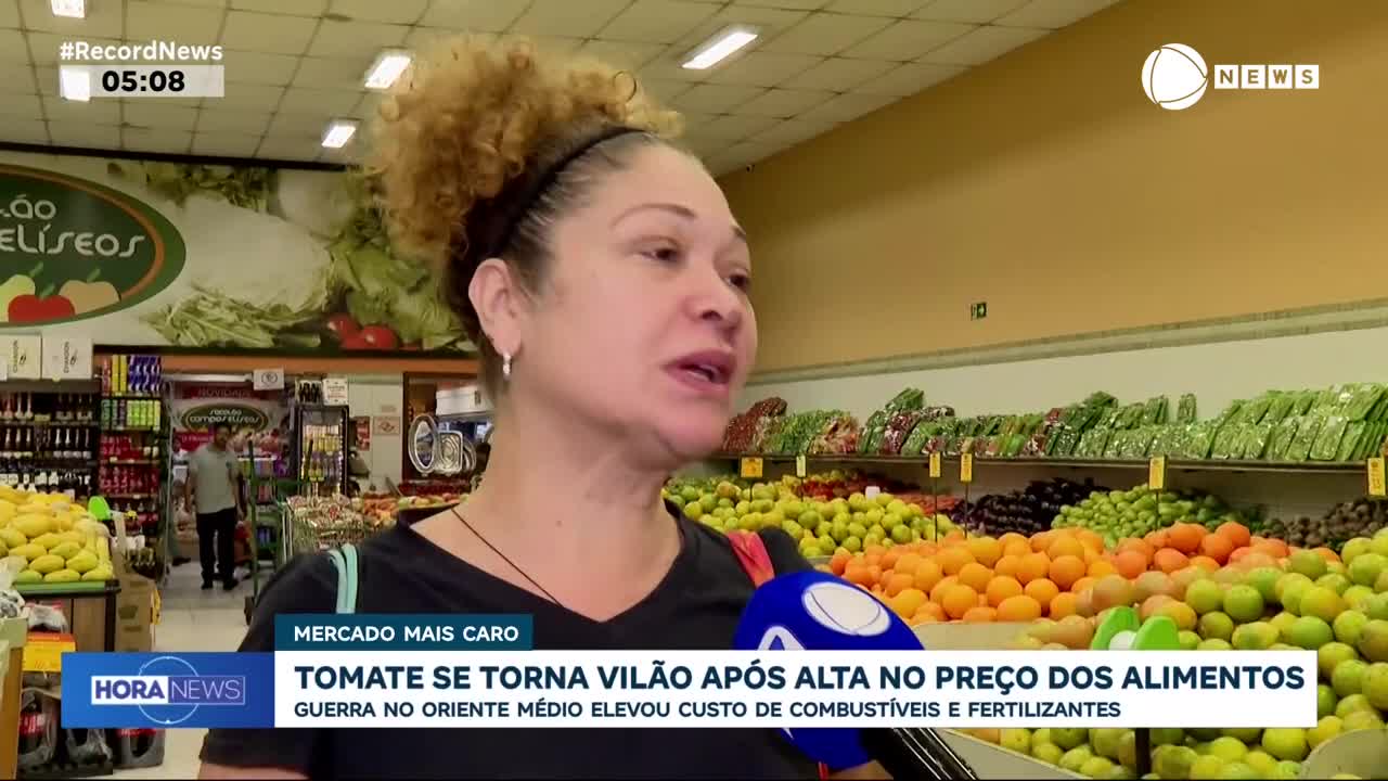 A woman is speaking into a microphone at a grocery store in Brazil. Behind her, shelves overflow with colorful fruits and vegetables.