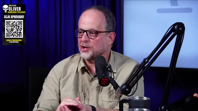 A man with glasses and a beard speaks into a microphone, his mouth open in mid-sentence. Behind him, a blue backdrop and a promotional graphic are visible.
