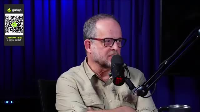 A man with glasses is speaking into a microphone, his face lit by the studio lights. Behind him, a blue curtain forms the backdrop for what looks like a Brazilian broadcast.
