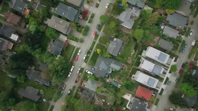 From this vantage, I see a grid of houses, each with a dark roof and surrounded by green trees. Cars are parked along the streets, and a few red ones stand out.
