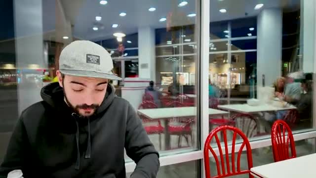 A man in a gray cap and black hoodie is looking down, seemingly focused on something in front of him. Through the window, other people are seated at tables in a brightly lit restaurant.
