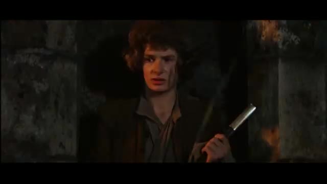 A young man, face streaked with dirt, stares directly ahead, holding a silver-colored weapon. He's framed by dark stone walls, lit by flickering flames.
