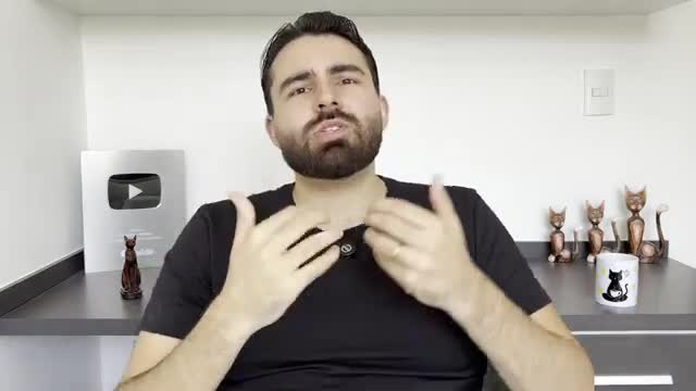 A man with a beard gestures with his hands as he speaks, presumably for a video. Behind him, a shelf holds cat figurines and a mug, along with a YouTube Play Button award.
