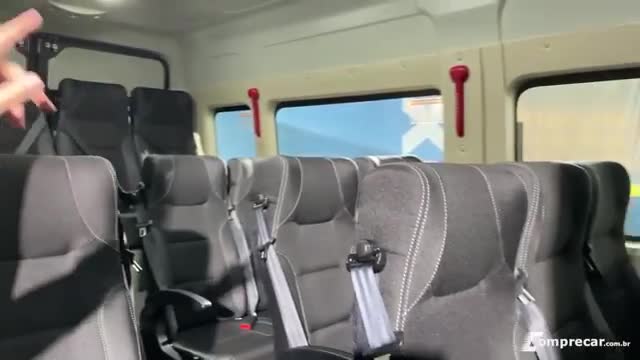 Inside a van, rows of dark gray seats face forward, each with a seatbelt buckled. A hand gestures, likely pointing out features of the vehicle's interior.
