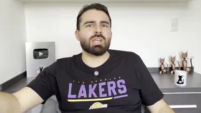 A man with a beard and a Lakers t-shirt is speaking, his mouth moving as he talks. Behind him, a silver YouTube play button sits on a shelf with cat figurines.
