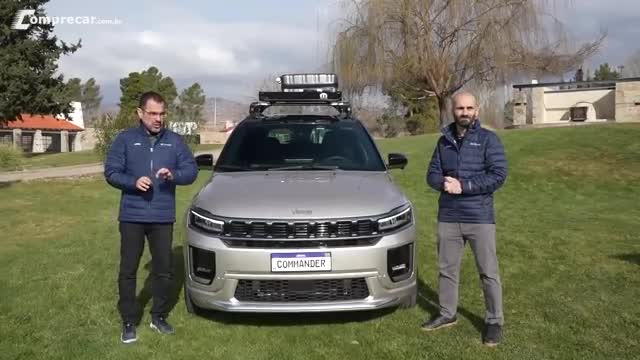 Two men in blue jackets stand on a grassy lawn, gesturing toward a silver Jeep Commander. The car's license plate reads "Commander," and it has a roof rack.
