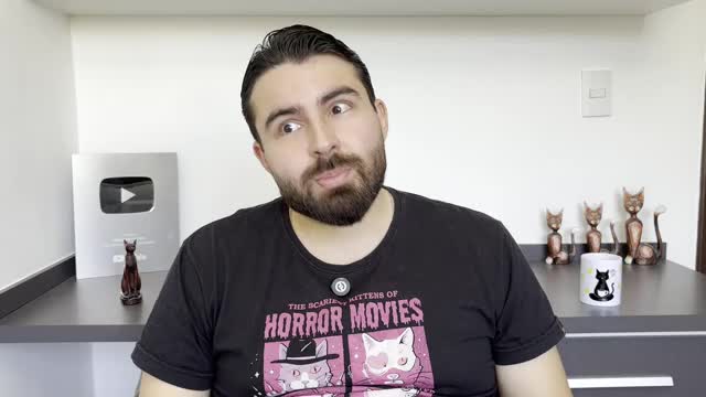 The man in the black t-shirt is looking off to the side, his eyes wide and his mouth slightly open. Behind him, a YouTube play button award and cat figurines sit on a dark surface.
