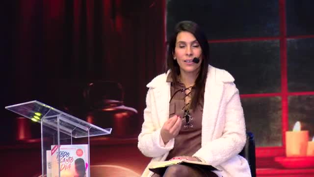 A woman in a white coat gestures with her hand while speaking into a microphone, likely on a Rede SPTV program. A book titled "Todo Santo Dia" rests on the podium next to her.
