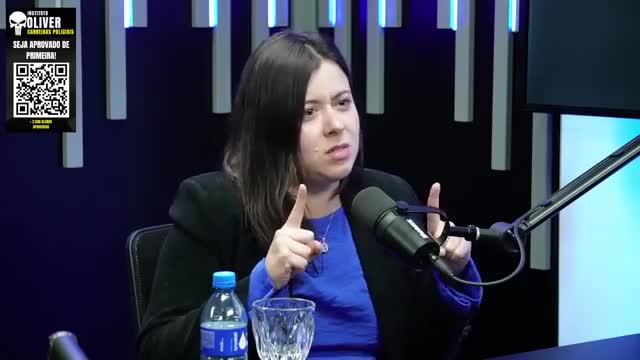 A woman in a blue shirt and black blazer gestures with both hands, pointing her index fingers. A microphone is positioned close to her face, and there's a bottle of water and a glass on the table in front of her.
