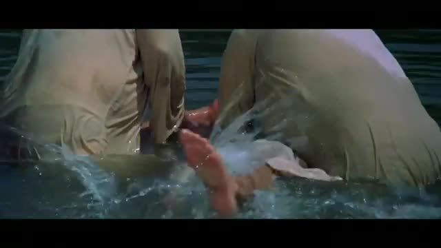 Two figures, clad in wet, pale clothing, are submerged in water, their legs and hands visible as they splash. The water churns around them, creating small waves and droplets.
