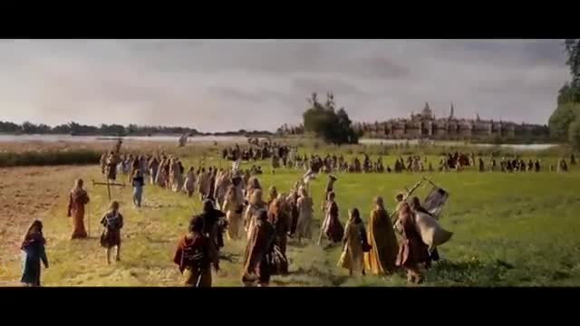 A long procession of people in simple clothing moves across a field toward a distant building. Some carry crosses and banners as they walk, and the group seems to be heading toward a large structure in the distance.
