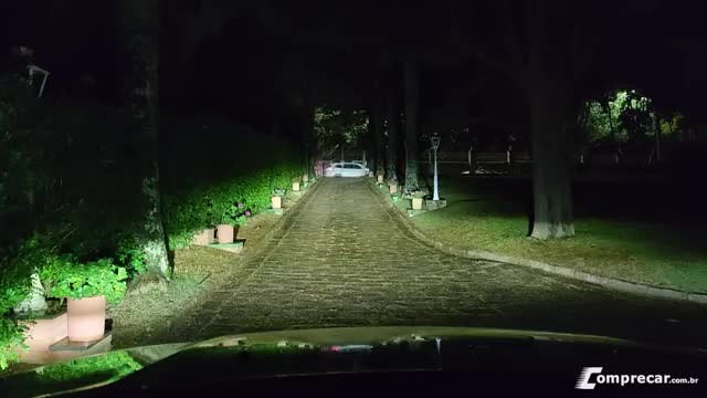 Headlights illuminate a long, brick driveway lined with trees and hedges. A white car sits at the end, bathed in the glow of the lights.
