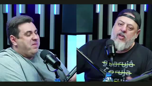 Two men are seated, each with a microphone in front of them. The man on the right, wearing a black cap and tee, is speaking animatedly.
