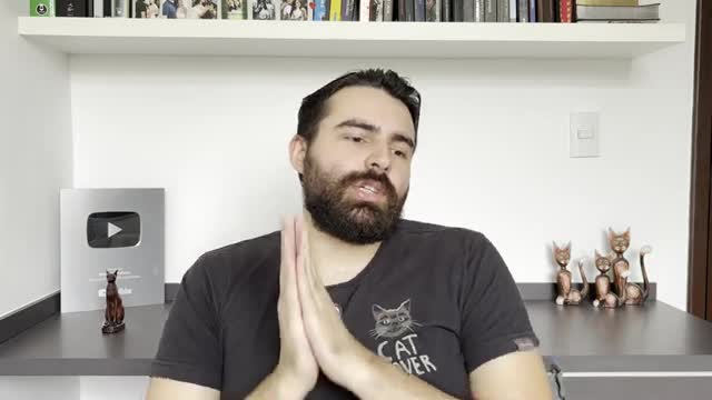 A man with a full beard clasps his hands together, looking directly at the camera. He's wearing a black t-shirt with a cat design, and behind him, a shelf holds books and decorative cat figurines.
