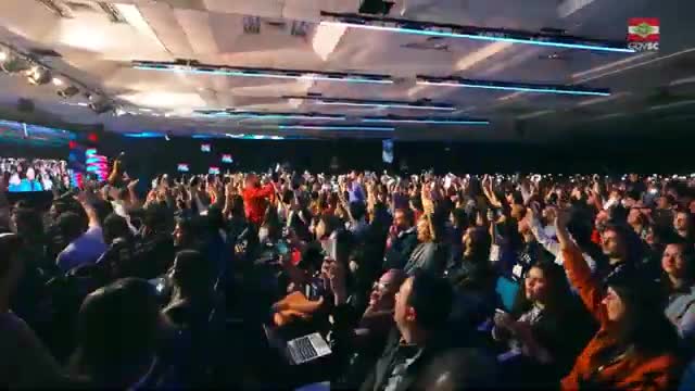 Hundreds of people in a large room are raising their arms, many holding phones. The energy is high, with the crowd's excitement palpable.
