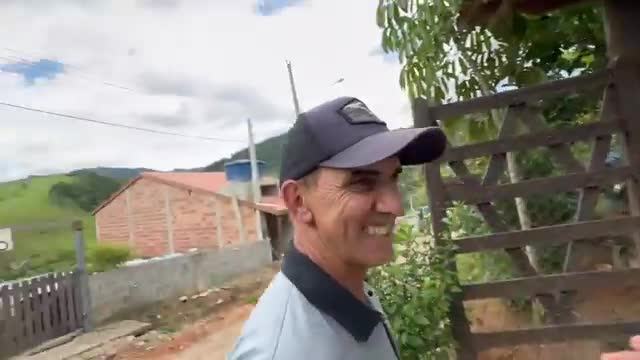 The man in the cap grins, turning his head slightly to the right. Behind him, a wooden gate and a brick house sit against a backdrop of green hills.
