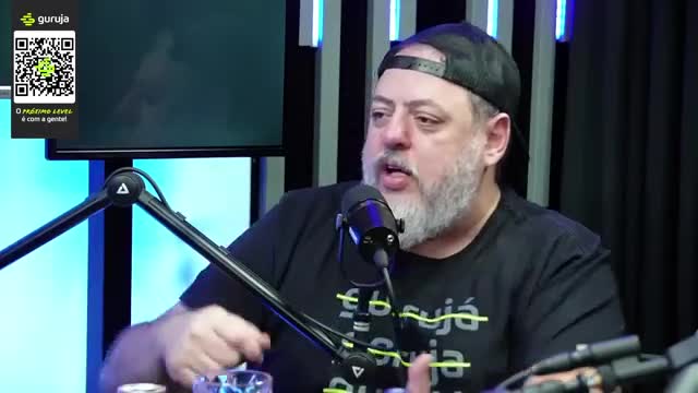 The man in the black cap gestures with his right hand while speaking into a microphone. A QR code for "Guruja" is displayed on a screen behind him.
