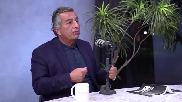 The man in the dark blazer gestures with his hands, speaking into a microphone. A white mug sits on the table in front of him, and a book lies open nearby.
