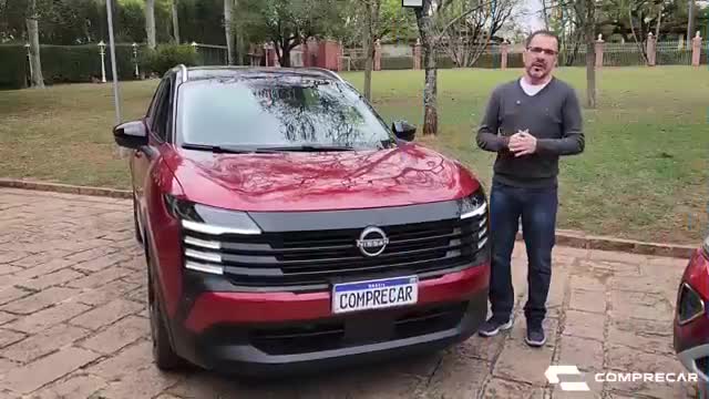 A man in glasses stands next to a shiny, red car, gesturing with his hands. The car's front grill and headlights are prominently displayed, with a "Comprecar" license plate visible.
A man in glasses stands next to a shiny, red car, gesturing with his hands. The car's front grill and headlights are prominently displayed, with a "Comprecar" license plate visible.