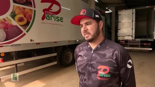 A man in a cap and camo shirt speaks next to a truck plastered with fruit imagery. Behind him, an empty trailer waits in a loading bay.
A man in a cap and camo shirt speaks next to a truck plastered with fruit imagery. Behind him, an empty trailer waits in a loading bay.