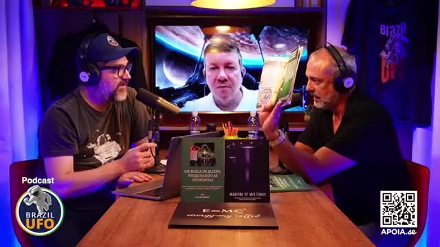 Two men wearing headsets sit at a table, microphones between them, with a screen showing a third person. The man on the right holds a book open, gesturing with it.
Two men wearing headsets sit at a table, microphones between them, with a screen showing a third person. The man on the right holds a book open, gesturing with it.