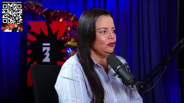 A woman with dark hair speaks into a microphone, her gaze directed off-camera. Behind her, a red and black backdrop is visible, along with a small inset screen featuring two men.
A woman with dark hair speaks into a microphone, her gaze directed off-camera. Behind her, a red and black backdrop is visible, along with a small inset screen featuring two men.