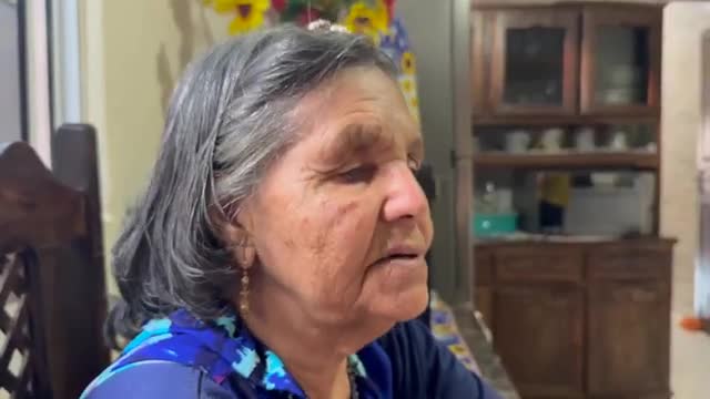 An older woman with gray hair is speaking, her mouth slightly open. She's wearing a patterned blue top, and a wooden cabinet sits behind her.
An older woman with gray hair is speaking, her mouth slightly open. She's wearing a patterned blue top, and a wooden cabinet sits behind her.