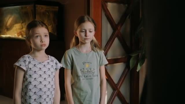 Two young girls stand near a doorway, one in a white shirt with a geometric pattern, the other in a light green t-shirt. The girl on the left looks directly ahead with a serious expression, while the one on the right gazes downward.