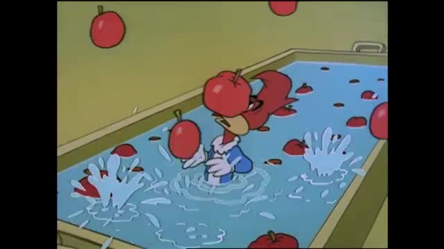 Apples are splashing into a tub of water as a red bird, looking like he's on Rede SPTV, reaches for one. The bird, wearing a blue and white outfit, is partially submerged in the water.