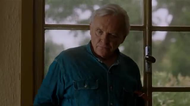 The man in the blue shirt looks out the window, his gaze fixed on something beyond the glass. A faint smile plays on his lips as he seems lost in thought.