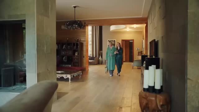 Two women walk down a long hallway, one in a green robe, the other in a dark teal outfit. The scene appears to be from a Brazilian production, perhaps for Rede SPTV.