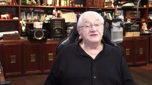 An older gentleman with white hair and glasses sits in a gaming chair, speaking into a microphone. Behind him, shelves are filled with books and vintage electronics, hinting at a studio setup for Rede SPTV.